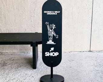 May include: A black vertical oval sign on a circular base featuring the white text GEORGE & WILLY ORGANICS, a white line drawing of a person in a hat and plaid shirt, an upward-pointing arrow, and the white text SHOP at the bottom.