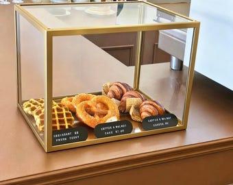 May include: A rectangular glass display case with a gold metal frame containing waffles, a pretzel, and croissants. Three black labels are placed in front of the pastries with white text reading "CROISSANT $8 FRESH TODAY", "COFFEE & WALNUT CAKE $7.00", and "COFFEE & WALNUT CAKE $8.00".