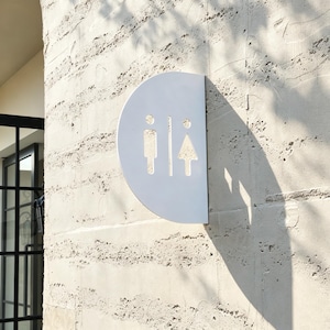 May include: A semi-circular white metal restroom sign mounted on a textured light-colored wall. The sign features cut-out silhouettes of a person in a rectangular shape and a person in a triangular shape, separated by a vertical line.