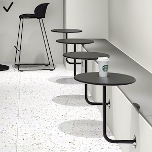 Peut inclure: Intérieur moderne avec un tabouret de bar noir et plusieurs petites tables rondes noires fixées à un mur blanc. Un gobelet en papier blanc avec un logo vert est posé sur une table. Le sol est blanc avec un motif moucheté.
