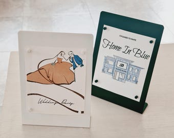 May include: Two rectangular metal sign holders, one white and one dark green, each displaying a printed card. The white holder features an illustration of a couple and the text Wedding Party. The green holder shows a building and the text Home In Blue.