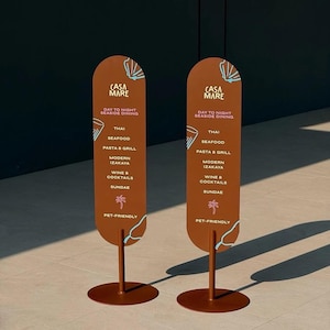 Può includere: Due insegne di ristoranti in metallo marrone con la scritta "CASA MARE" e un elenco di voci di menu tra cui Thai, frutti di mare e vini e cocktail. I cartelli sono di forma ovale con una grafica di palma e sono adatti agli animali.