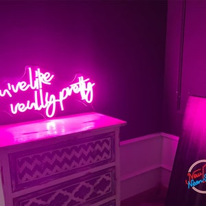 May include: A pink neon sign with the words "You've like really pretty" in a cursive script. The sign is positioned above a white, distressed wooden chest of drawers. The background is a dark purple wall.