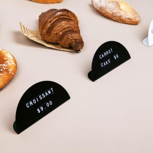 May include: Two semi-circular black metal display stands featuring white embossed text. One sign reads CROISSANT $9.00 and the other reads CARROT CAKE $6. The stands are positioned near a golden-brown croissant on a piece of printed paper.