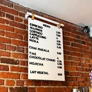 Op de afbeelding: Een wit menubord met zwarte letters hangt aan een witte balk met leren riemen tegen een rode bakstenen muur. Het menu bevat koffie- en theedranken, waaronder Espresso en Latte.