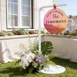 Puede incluir: Un letrero de boda de metal blanco con un letrero circular que dice "Welcome to our Wedding the Ceremony" en texto negro. El letrero es rosa y naranja con los nombres Alice y Mason y la fecha 17 de septiembre de 2026. El letrero está rodeado de flores.