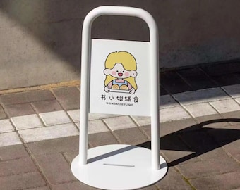 May include: A white metal sign with a circular base and a curved top handle. The sign features a cartoon illustration of a girl with blonde hair holding baked goods, with text in Chinese characters below.
