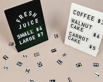 May include: A black sign with white text reading FRESH JUICE, SMALL $4, LARGE $7 and a white sign with black text reading COFFEE $3, WALNUT CAKE $6, CARROT CAKE $5. Scattered individual black and white letter and number tiles surround the two signs.