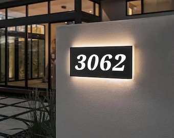 May include: A rectangular black address sign with the number "3062" illuminated in white. The sign is mounted on a light-colored wall, with a warm glow emanating from behind the numbers.