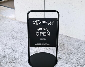 May include: A black metal A-frame sidewalk sign with a round base. The sign features white text that reads: minon, we are OPEN, wed-mon 8am-10pm, and come on in for some warm coffee and tasty treats! The frame is constructed from thin black tubing.