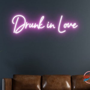 May include: Pink neon sign with the text "Drunk in Love" mounted on a dark gray wall above a brown leather couch.