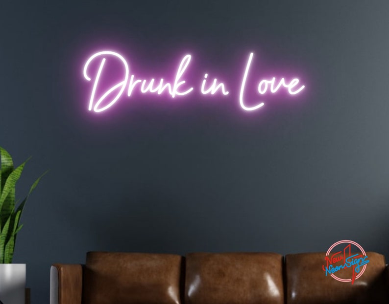 May include: Pink neon sign with the text "Drunk in Love" mounted on a dark gray wall above a brown leather couch.