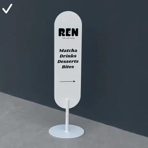 May include: A white vertical oval sign on a circular base stand. The sign features black text reading REN Cafe and Goods, followed by Matcha, Drinks, Desserts, Bites, and a right-pointing arrow. The design is clean, featuring bold black lettering.