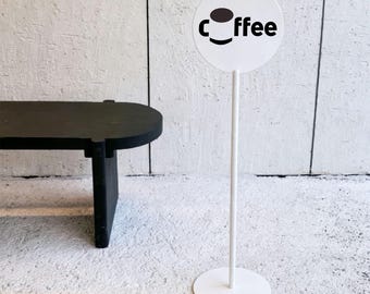 May include: A white circular sign on a thin vertical stand with a round base. The sign features the word coffee in black lowercase letters, with the letter o replaced by a graphic of a brown and white cup. The stand and base are white.