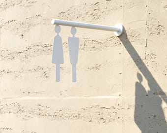 May include: A white metal wall-mounted sign featuring two stylized human figures hanging from a horizontal bar. The figures represent a person in a dress and a person with a rectangular torso, both suspended by their heads from the white rod.
