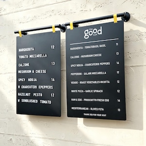 May include: Two black rectangular menu boards hanging from a black metal rod with yellow clips. The boards display food items and prices in white text, including "THE GOOD LIFE" at the top, listing various pizzas like Margherita, Calzone, and Nduja.