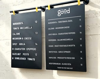 May include: Two black rectangular menu boards hanging from a black metal rod with yellow clips. The boards display food items and prices in white text, including "THE GOOD LIFE" at the top, listing various pizzas like Margherita, Calzone, and Nduja.