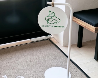 May include: A white metal floor sign featuring a circular panel suspended from a curved stand. The panel displays a green line drawing of a person wearing a hat with a small bird on top, accompanied by the green text EGG IN THE MORNING.