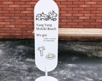 May include: A white oval-shaped standing sign with a black graphic of waves and the text Kinship, Yang Yang Mulchi Beach, We got Coffee, Cocktail & Community, featuring line drawings of a palm tree and a slice of cake on a plate.
