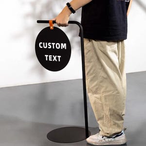 Puede incluir: Un letrero de metal negro con un área de visualización circular. El letrero dice "CUSTOM TEXT" en letras blancas. El letrero está sostenido por un soporte de metal negro con una base redonda y una correa de cuero naranja.