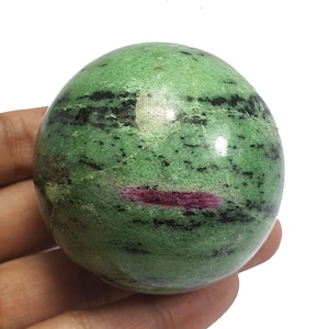 Puede incluir: Una esfera pulida de zoisita verde con inclusiones negras y rojas. La gema se sostiene en una mano sobre un fondo blanco. La superficie de la esfera tiene un acabado liso y brillante, resaltando los patrones naturales.