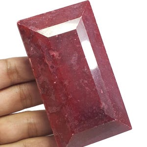 Puede incluir: Una gran piedra preciosa de rubí rectangular con un tono rojo intenso. La gema tiene un corte facetado con ángulos agudos y una superficie pulida, que refleja la luz. El rubí se sostiene en una mano sobre un fondo blanco.