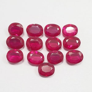 May include: An assortment of oval-cut, deep red rubies. The gemstones are arranged in a grid pattern, showcasing their faceted surfaces and vibrant color. The rubies are set against a plain white background.