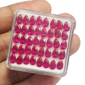 May include: A clear plastic case filled with 36 vibrant, teardrop-shaped, faceted rubies. The gems are a deep, rich pink-red color, and are arranged neatly in rows. The case is held in a hand.