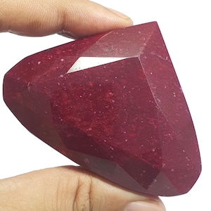 May include: A large, faceted, deep red gemstone with a triangular shape. The stone features a polished surface with visible internal inclusions and a vibrant, saturated crimson color throughout its structure, showcasing multiple flat geometric faces.