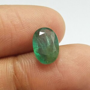 May include: Oval-cut emerald gemstone. The gem is a vibrant green colour with visible inclusions and facets. The stone is held between two fingers against a white background, showcasing its clarity and brilliance.