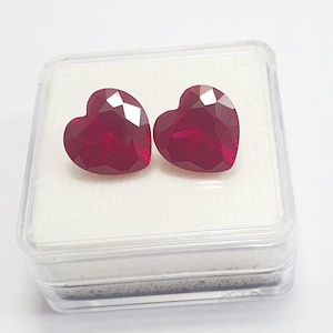 Könnte beinhalten: Zwei herzförmige, facettierte, tiefrote Edelsteine werden in einer transparenten Kunststoffbox mit einem weißen Schaumstoffeinsatz präsentiert. Die Edelsteine haben mehrere Facetten, die das Licht einfangen und einen funkelnden Effekt erzeugen.