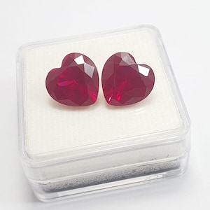 May include: Two heart-shaped, faceted, deep red gemstones are displayed inside a clear plastic case. The gems have multiple facets that catch the light, creating a sparkling effect. The case sits on a white surface.