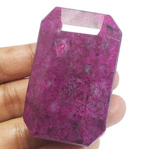 May include: A rectangular, faceted gemstone pendant in a deep magenta-pink hue with natural inclusions and a flat top edge featuring a horizontal rectangular cutout for suspension. The stone has a polished surface with visible internal texture.