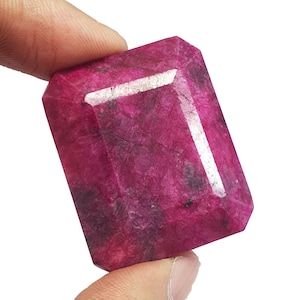 Könnte beinhalten: Ein facettierter, rechteckiger Rubin-Edelstein in tiefroter Farbe. Der polierte Edelstein hat sichtbare innere Einschlüsse und eine helle, reflektierende Facette. Der Rubin wird zwischen zwei Fingern vor weißem Hintergrund gehalten.