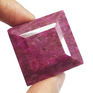 May include: A square-cut, faceted gemstone in shades of deep pink and red. The gem is held between fingers, showcasing its polished surface and geometric design. The gemstone's colour is rich and vibrant, with subtle variations in tone.