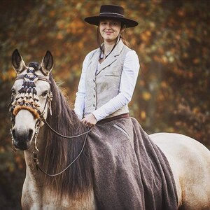 Puede incluir: Una mujer con una camisa blanca y un chaleco marr&oacute;n monta un caballo con una brida de cuero marr&oacute;n y un frontal decorativo. El caballo es blanco con una crin y una cola marrones. La mujer lleva un sombrero negro y una falda marr&oacute;n. El fondo es una imagen borrosa de &aacute;rboles.
