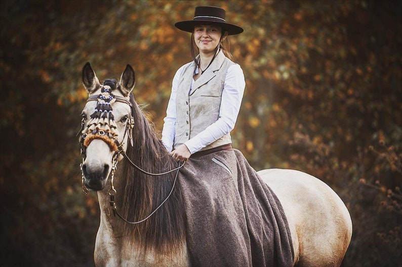 Puede incluir: Una mujer con una camisa blanca y un chaleco marr&oacute;n monta un caballo con una brida de cuero marr&oacute;n y un frontal decorativo. El caballo es blanco con una crin y una cola marrones. La mujer lleva un sombrero negro y una falda marr&oacute;n. El fondo es una imagen borrosa de &aacute;rboles.
