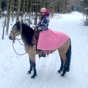 Puede incluir: Una persona con un casco rosa y una chaqueta floral monta un caballo marr&oacute;n y negro en un bosque nevado. El caballo lleva una manta de silla rosa.
