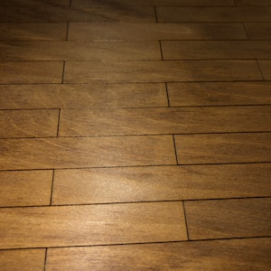 May include: Close-up of a wooden floor with a warm, brown tone. The floor is made of rectangular planks arranged in a parallel pattern. The wood grain is visible, adding texture and detail to the surface. The planks are separated by thin, dark lines.