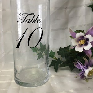 May include: A clear glass cylinder vase with the words "Table 10" in black script. The vase is surrounded by greenery and purple and white flowers, suggesting a wedding or event theme.