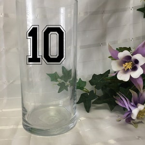 May include: A clear glass cylinder vase with the number 10 in black with a white outline. The vase is decorated with green ivy and purple and white flowers.