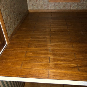 May include: A miniature dollhouse interior featuring a wooden floor with a warm, brown stain. The floorboards are rectangular and arranged in a staggered pattern. The walls are adorned with patterned wallpaper, and a window is visible in the background.