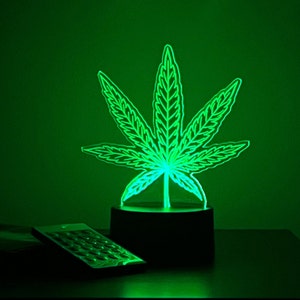May include: A green LED night light shaped like a cannabis leaf. The light sits on a black base, with a remote control in the foreground. The background is a solid green.
