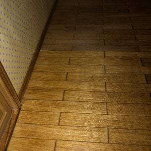 May include: A miniature wooden floor with a warm, brown finish. The planks are arranged in a staggered pattern, creating a realistic look. The floor is next to a wall with yellow wallpaper and a decorative border.