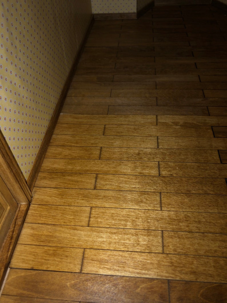 May include: A miniature wooden floor with a warm, brown finish. The planks are arranged in a staggered pattern, creating a realistic look. The floor is next to a wall with yellow wallpaper and a decorative border.