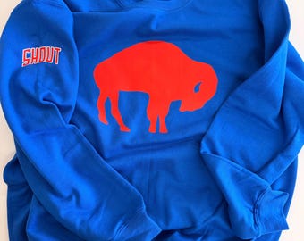 Puede incluir: Una sudadera de cuello redondo azul real que presenta una gran silueta de búfalo roja sólida centrada en el pecho. La palabra SHOUT está impresa en blanco con un contorno rojo en la manga derecha. La prenda tiene mangas largas y cuello acanalado.