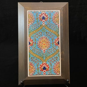 Op de afbeelding: Een ingelijste tegel met een kleurrijk bloemenpatroon in tinten rood, geel, blauw en groen. De tegel is geplaatst op een blauwe achtergrond en heeft ingewikkelde details.