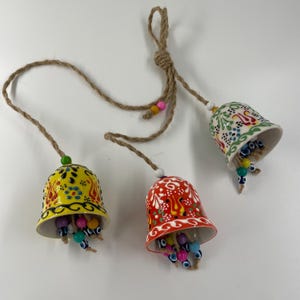 Puede incluir: Tres campanas de cerámica con coloridos patrones florales y cuentas azules de ojo turco colgando de una cuerda de yute. Las campanas son amarillas, rojas y blancas.