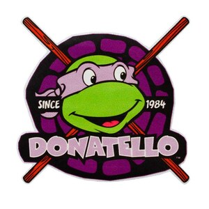 Könnte beinhalten: Ein Aufnäher mit Donatello von den Teenage Mutant Ninja Turtles. Das Design zeigt einen grünen Schildkrötenkopf mit violetter Maske, gekreuzten roten Bo-Stäben und den Texten "DONATELLO" und "SINCE 1984". Der Hintergrund ist lila und schwarz.