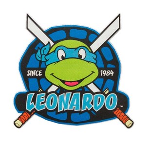 Könnte beinhalten: Ein farbenfrohes Grafikdesign mit Leonardo von den Teenage Mutant Ninja Turtles. Das Design zeigt ein grünes Schildkrötengesicht mit blauer Maske, gekreuzten weißen Schwertern und dem Namen "LEONARDO" in blauen Buchstaben. Der Text "SINCE 1984" ist ebenfalls enthalten.